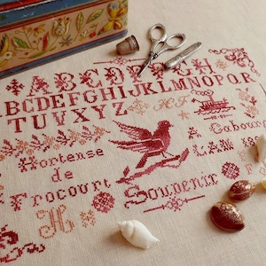 May include: A vintage cross-stitch sampler in red thread features the alphabet, a bird, and the words "Hortense de Frocourt Souvenir." The sampler is accompanied by seashells, a thimble, scissors, and a small container.