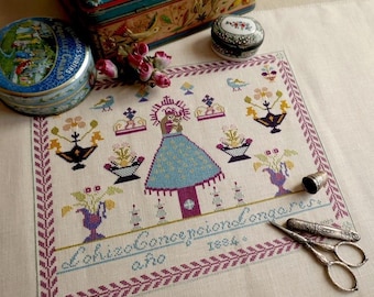 Pode incluir: Um bordado de ponto cruz detalhado com uma figura religiosa num vestido azul, rodeada por motivos florais e coroas. O desenho é emoldurado com uma borda roxa e inclui texto. Uma dedaleira e uma tesoura estão ao lado do bordado.