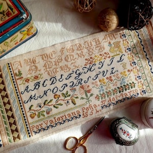 May include: A detailed cross-stitch sampler with an alphabet and floral motifs. The sampler features various colors of thread on a cream-colored fabric. The text "Por Valeriana Peña. 17 de Marzo" is stitched at the bottom. A vintage tin and scissors are also visible.
