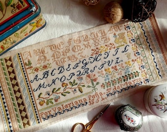 May include: A detailed cross-stitch sampler with an alphabet and floral motifs. The sampler features various colors of thread on a cream-colored fabric. The text "Por Valeriana Peña. 17 de Marzo" is stitched at the bottom. A vintage tin and scissors are also visible.