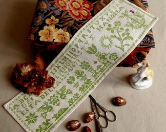 Pode incluir: Um bordado em ponto cruz de linho branco com bordados verdes, com letras, números e motivos florais. O bordado está ao lado de uma caixa com estampa floral, uma pequena figura de cão e ovos decorativos. Também estão presentes um par de tesouras e uma flor seca.