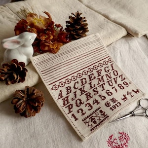 May include: A vintage-style cross-stitch sampler with the alphabet and numbers in burgundy thread. The sampler is on a cream-colored fabric, with decorative borders and the year 1913. A white ceramic bunny, pine cones, and scissors are nearby.