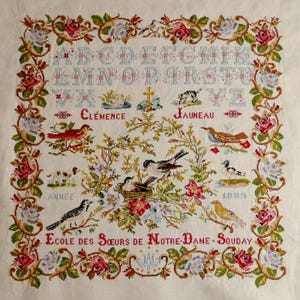 Peut inclure: Une broderie au point de croix vintage avec un alphabet, des oiseaux et des motifs floraux. Le motif comprend les noms "Clémence" et "Jauneau", et le texte "Ecole des Sœurs de Notre-Dame-Souday". La bordure est ornée de roses et d'autres fleurs.