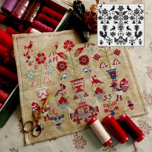 May include: A cross-stitch sampler featuring red, blue, and white floral and bird designs on a beige linen fabric. Gold scissors and red thread spools are placed near the sampler. A black and white chart is in the upper right corner.