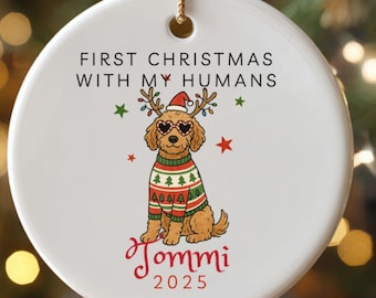May include: White ceramic Christmas ornament with a gold string. The ornament features a cartoon dog wearing a Christmas sweater and sunglasses. Text reads "First Christmas with my humans" and "Tommi 2025".