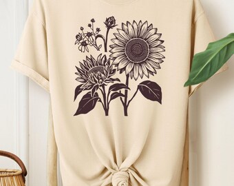 May include: Ivory-colored t-shirt featuring a detailed black and white floral design of sunflowers and other flowers. The shirt is knotted at the waist and hangs on a wooden rack. The word "IVORY" is in a pink box.