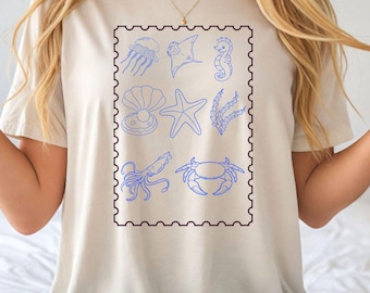May include: Ivory-colored t-shirt featuring a blue stamp design with various sea creatures. The stamp includes a jellyfish, stingray, seahorse, seashell, starfish, seaweed, octopus, and crab. The word "IVORY" is printed in red.
