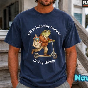 Peut inclure: Un t-shirt bleu marine avec une illustration d'une grenouille portant des lunettes et une veste marron, sur une trottinette avec un sac fourre-tout rempli de fournitures scolaires. Le texte "Off to help tiny humans do big things" est en blanc.