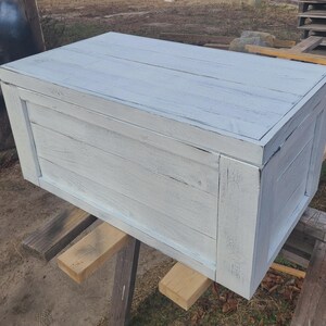May include: A rectangular wooden storage chest painted in a distressed white finish. The chest is constructed with horizontal planks and framed with vertical supports. The wood grain is visible through the paint.