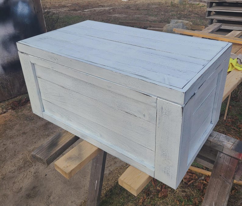 May include: A rectangular wooden storage chest painted in a distressed white finish. The chest is constructed with horizontal planks and framed with vertical supports. The wood grain is visible through the paint.