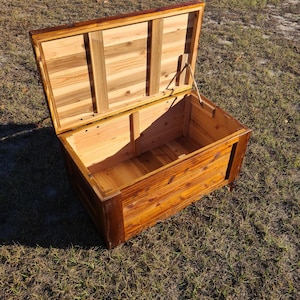 May include: Open wooden chest with a hinged lid. The chest is made of light and dark brown wood, with a rectangular shape. The lid is propped open, revealing the interior. The chest is sitting on a grassy surface.