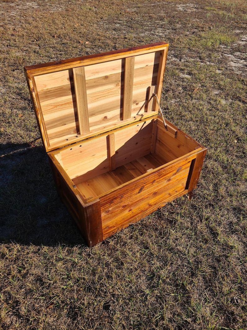 May include: Open wooden chest with a hinged lid. The chest is made of light and dark brown wood, with a rectangular shape. The lid is propped open, revealing the interior. The chest is sitting on a grassy surface.