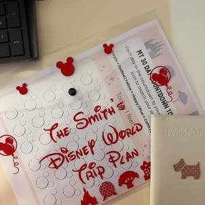 May include: A clear plastic sleeve with a red Disney World Trip Plan, Mickey Mouse-shaped embellishments, and a passport cover. The text on the plan reads "The Smith's Disney World Trip Plan". The passport cover is off-white with a dog graphic.