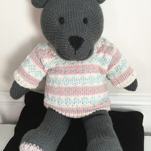 May include: A grey knitted teddy bear wearing a pink and blue striped jumper. The bear is sitting on a black cushion.