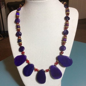 May include: A purple and red beaded necklace with large purple gemstone pendants. The necklace is displayed on a white mannequin bust.