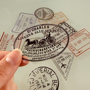 Op de afbeelding: Transparante stickers in vintage-stijl met diverse ontwerpen met een reisthema. Eén sticker toont een paard en wagen en de tekst "St. Charles Livery & Sale Staples". Andere stickers bevatten stempels met data en locaties.