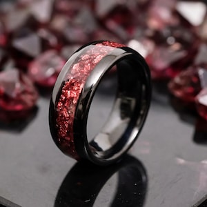 May include: A black band ring featuring a central inlay of crushed red stone fragments. The ring has a polished, reflective surface and a smooth, rounded profile. The red inlay creates a textured, sparkling contrast against the dark metal band.