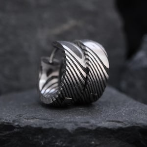 May include: A pair of silver hoop earrings with a unique striped pattern. The earrings are made of a shiny metal and have a textured design. The earrings are displayed on a dark gray surface.