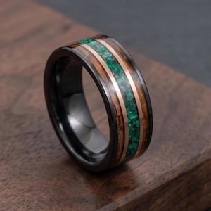 May include: A black ring featuring a central inlay of crushed green stone flanked by two thin copper-coloured metal bands and two outer bands of dark brown wood grain, all set within a polished black metal band with a smooth, rounded exterior profile.