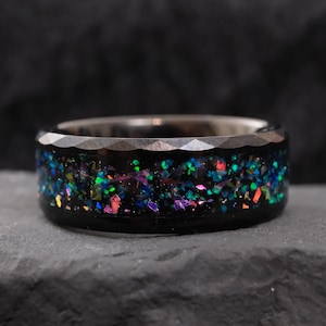 Puede incluir: Un anillo de carburo de tungsteno negro con un borde biselado y una banda de purpurina iridiscente incrustada en la resina negra.
