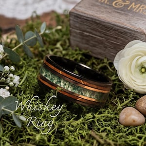 May include: A black ring with a green and brown inlay, with the text "Whiskey Ring" in white script. The ring is set on a bed of green moss, with a wooden box and white flowers in the background.