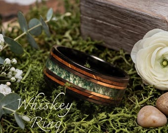 May include: A black ring with a green and brown inlay, with the text "Whiskey Ring" in white script. The ring is set on a bed of green moss, with a wooden box and white flowers in the background.