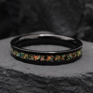 May include: A black ring with a band of colorful, iridescent stones. The ring is set on a dark, textured surface. The stones display hues of orange, green, and yellow.