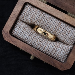 Peut inclure: Une alliance en or est posée dans un écrin en bois garni de toile de jute beige. La bague est très polie, reflétant l'environnement. Le coffret est ouvert, révélant la place de la bague.