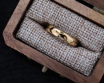 May include: A gold wedding ring sits in a wooden ring box lined with beige burlap. The ring is highly polished, reflecting the surroundings. The box is open, displaying the ring's position.