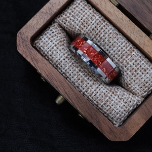 May include: A silver ring with a red and black speckled center band, flanked by silver and black stripes. The ring is displayed inside a small, open wooden box lined with tan fabric.