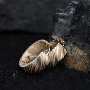 May include: Close-up of a gold and silver ring with a striped pattern. The ring is made of metal and has a unique design. The ring is resting on a black surface next to a dark gray rock.