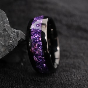 Op de afbeelding: Een zwarte ring met een band van fijngemaakte paarse edelstenen. De ring heeft een gepolijst, reflecterend oppervlak en de edelstenen zijn een mix van lichte en donkere paarse tinten. De ring staat tegen een donkere achtergrond.