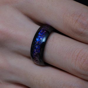 Könnte beinhalten: Ein schwarzer Ring mit einem Band aus blauem und violettem schillerndem Material. Der Ring befindet sich an einem Finger und zeigt die polierte, reflektierende Oberfläche und die leuchtenden Farben des eingelegten Designs. Der Ring ist ein einzigartiges Schmuckstück.