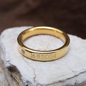 Peut inclure: Une bague dorée avec un petit diamant et la date gravée "16.9.2024". La bague est posée sur une surface en pierre blanche, mettant en valeur sa finition polie. La bague est de conception simple.