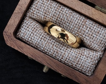 May include: A gold-colored ring with a polished surface rests inside a wooden ring box. The ring is set against a beige fabric lining. The box is open, revealing the ring's reflective surface.