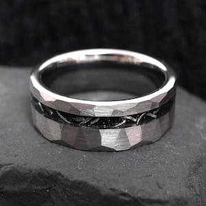 Könnte beinhalten: Ein silberfarbener Metallring mit einer gehämmerten, facettierten Textur an den oberen und unteren Kanten. Die Mitte des Bandes enthält eine vertiefte, tiefschwarze Einlage mit einem feinen, unregelmäßigen Metalldrahtmuster, das horizontal verläuft.