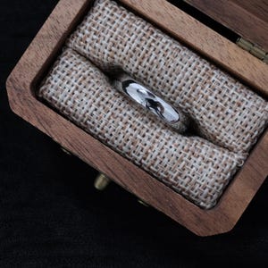 May include: A silver ring with a polished finish sits in a small wooden box lined with tan fabric. The ring reflects the surroundings. The box is open, showing the ring's position inside.