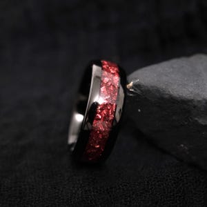 Op de afbeelding: Een zwarte ring met een centrale inleg van verbrijzelde rode steenfragmenten. De band heeft een gepolijst, reflecterend oppervlak dat contrasteert met het getextureerde, sprankelende uiterlijk van de rode inleg rondom de ring.