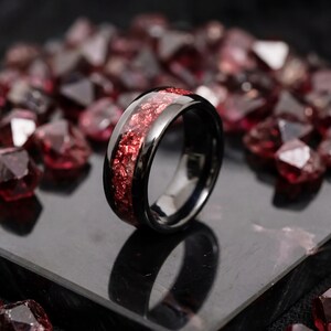 May include: A black ring with a polished finish, featuring a band filled with red, textured material. The ring is set against a backdrop of scattered red gemstones, creating a contrast of textures and colors.