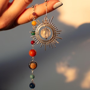 May include: Silver earrings with a sun design and a clear crystal. The earrings have a dangling chain with colorful beads representing planets.