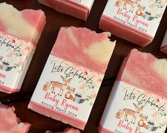 May include: Pink and white swirled soap bars with decorative labels. The labels read "Let's Celebrate Baby Ryann Arriving March 2024" and feature illustrations of woodland animals. The soaps are arranged on a dark wooden surface.