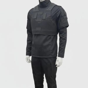 Puede incluir: Un atuendo táctico negro exhibido en un maniquí. El atuendo incluye una camisa de manga larga, un chaleco táctico y pantalones. El chaleco tiene múltiples bolsillos y paneles. Los pantalones tienen bolsillos en los lados y las rodillas. El atuendo está diseñado para protección y utilidad.