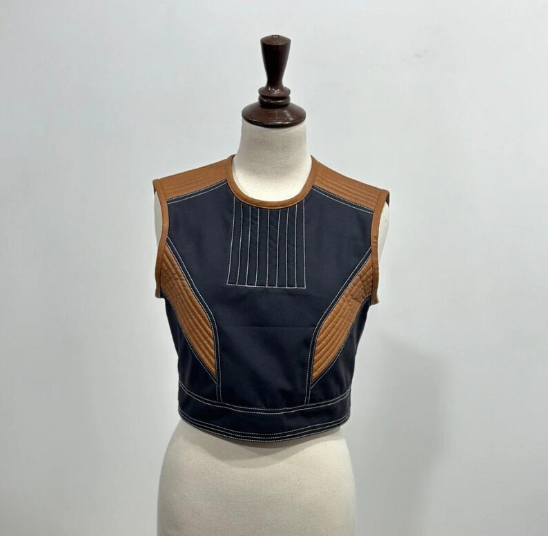 May include: A sleeveless, cropped top in black with brown accents. The top features quilted brown panels on the shoulders and sides, with white stitching details. Vertical white stitching adorns the center front.