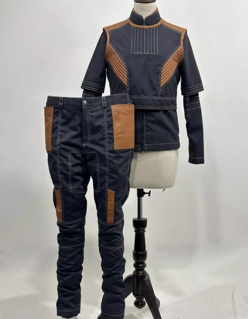 May include: A navy blue two-piece costume set, comprising a jacket and trousers. The jacket features brown shoulder and chest panels, with white stitching. The trousers have brown pockets and white stitching. The outfit is displayed on a mannequin.