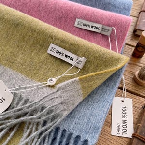 May include: A stack of three soft, textured scarves in light blue, pink, and a mustard yellow, gray, and white color-blocked pattern. Each scarf features a white tag with the text 100% WOOL and a small white circular tag with the letter W attached.