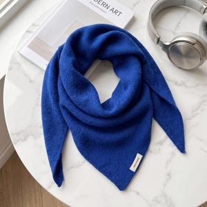 May include: A vibrant royal blue triangular scarf made of knit fabric. The scarf features a small rectangular white fabric tag sewn onto the bottom corner with the text 100% WOOL printed in black. The knit texture is visible throughout the piece.
