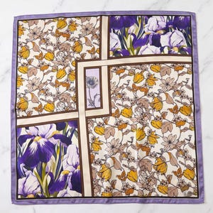 Op de afbeelding: Een vierkante sjaal met een gesegmenteerd ontwerp en een lichtpaarse rand. Het patroon bevat secties met gele en beige bloemenprints die afwisselen met panelen van paarse en witte irissen, gescheiden door dunne bruine lijnen.