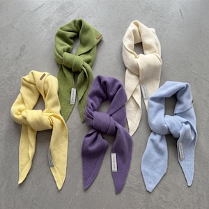 Puede incluir: Cinco bufandas de punto en amarillo pastel, verde oliva, morado, crema y azul claro. Cada bufanda está anudada y cuenta con una pequeña etiqueta de tela rectangular que dice 100% WOOL, indicando la composición del material de los accesorios.