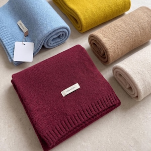 May include: A collection of five folded knit scarves in light blue, mustard yellow, tan, cream, and deep burgundy. Each scarf features a ribbed edge and a small white fabric tag with the text 100% WOOL. The light blue scarf includes an additional blank white tag.