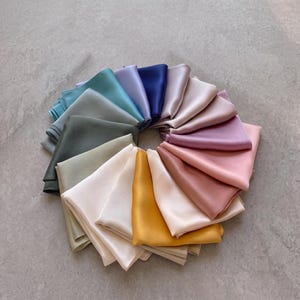 Op de afbeelding: Een collectie vierkante satijnen sjaals gerangschikt in een cirkelvormig waaierpatroon. De sjaals hebben een gladde, reflecterende afwerking en worden gepresenteerd in een gevarieerd palet met tinten blauw, groen, paars, roze, geel en crèmetinten.